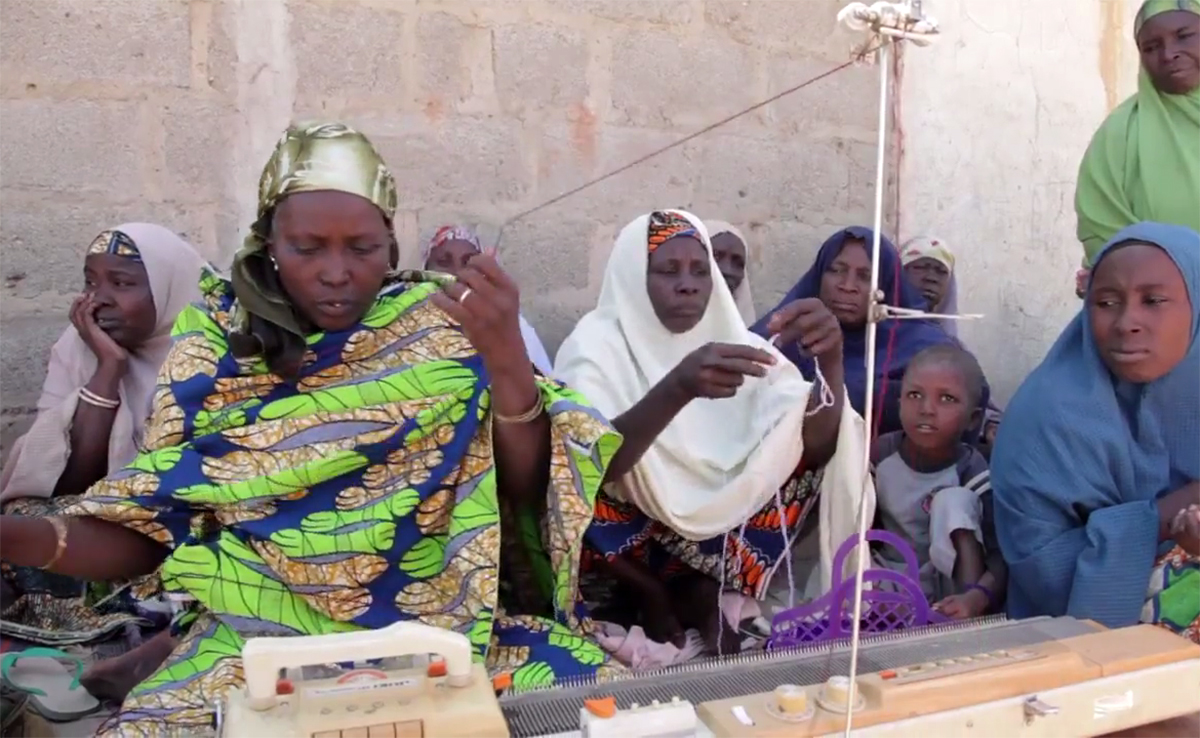 Women working on a loom outside with children and locals sitting beside them.