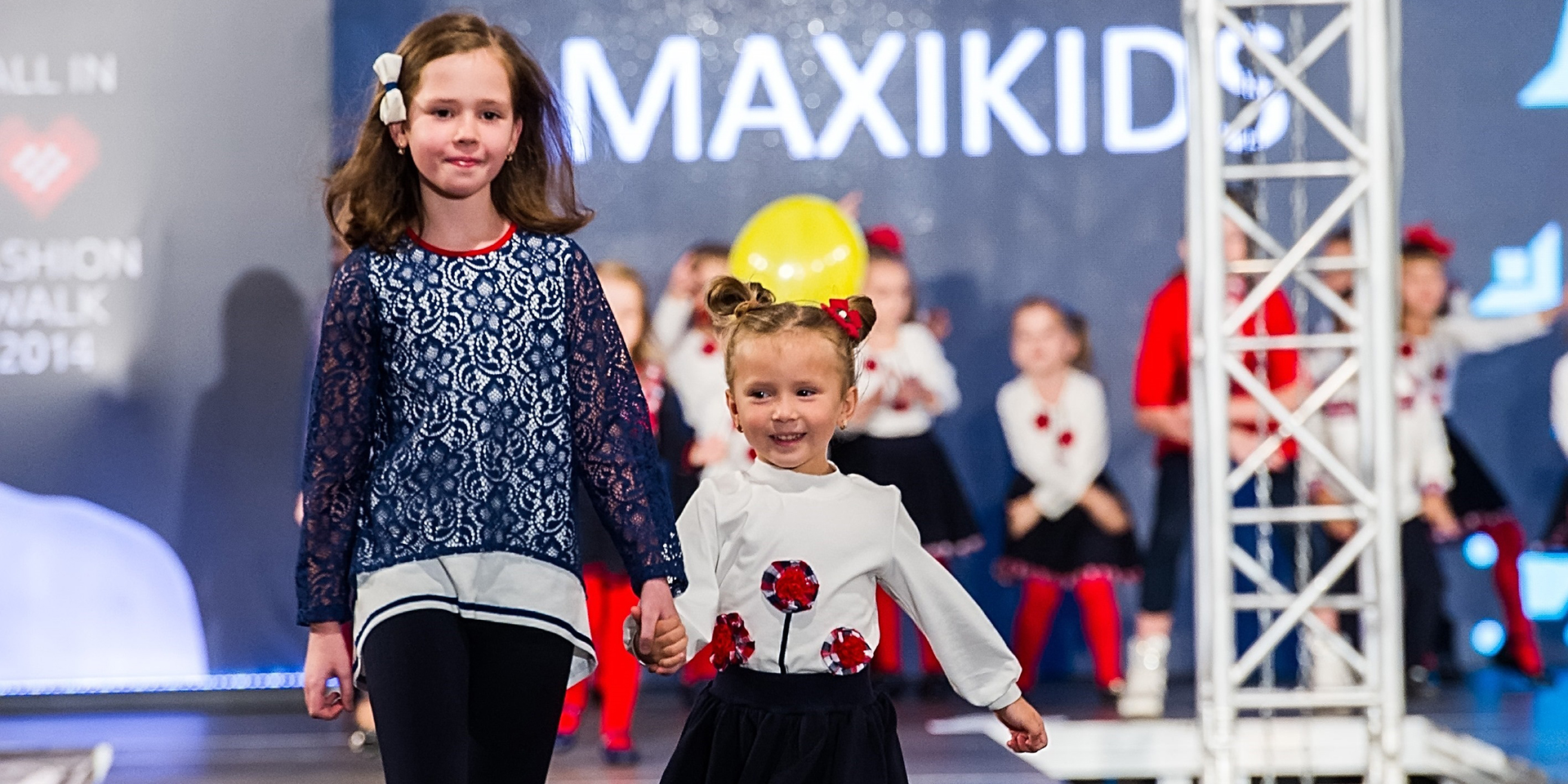 Two young girls smiling, holding hands, and walking down a fashion runway as other young girls wait their turn in the background.