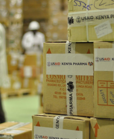 Image of a stack of boxes marked "Kenya Pharma" with several other stacks in the background being examined by warehouse workers.