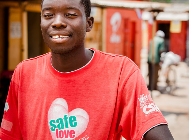 Image of a smiling young man on a busy street wearing a red t-shirt that says "Safe Love."