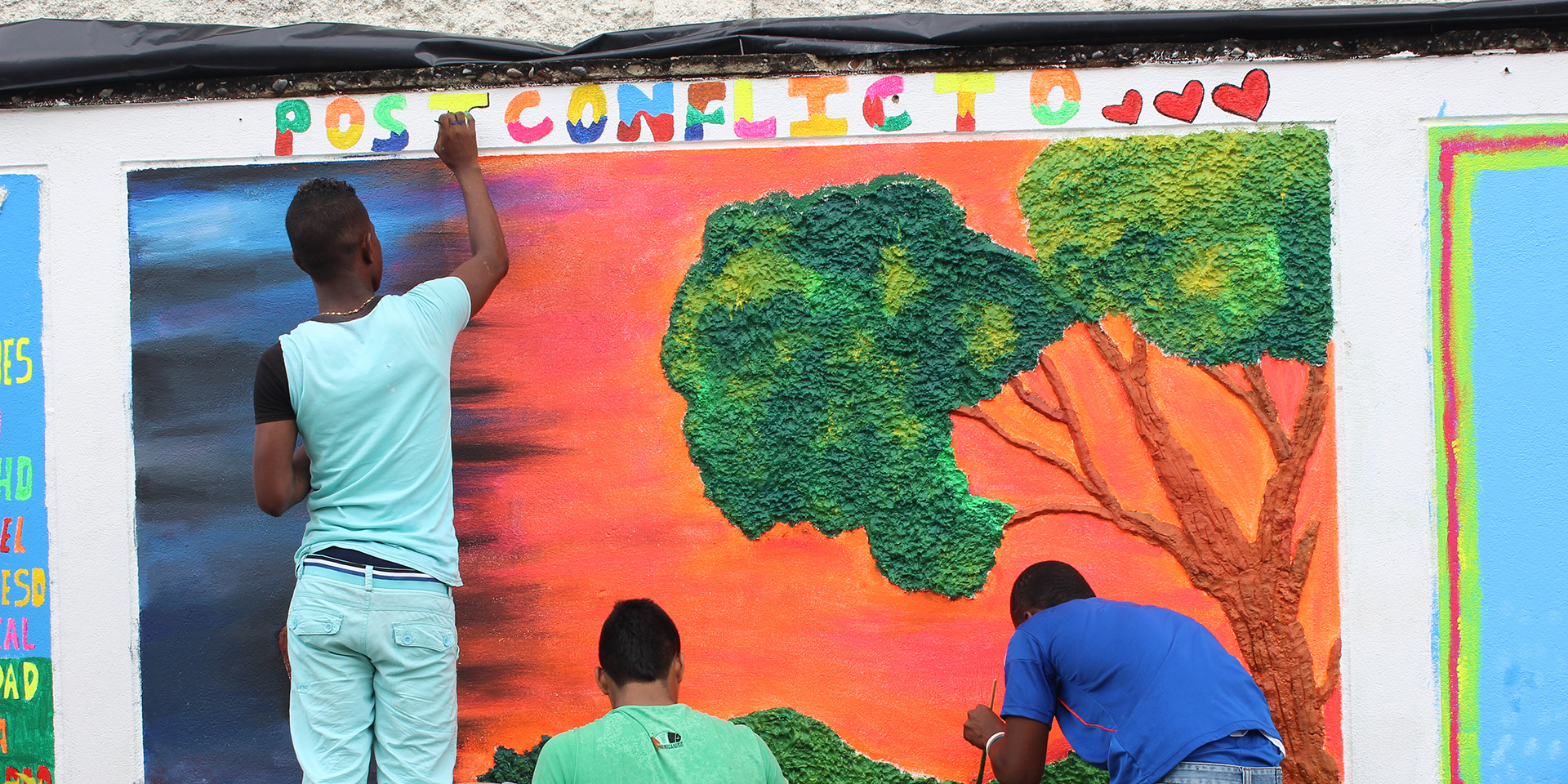 Three young men painting a vibrant mural of a tree with the word 