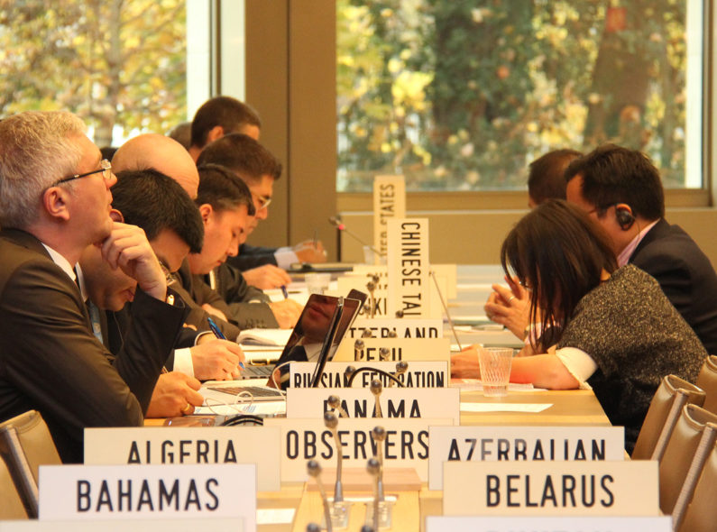 Several people sitting across from one another at a long table representing countries indicated by placards beside their seats, including "Russian Federation," "United States," and "Azerbaijan," among others.