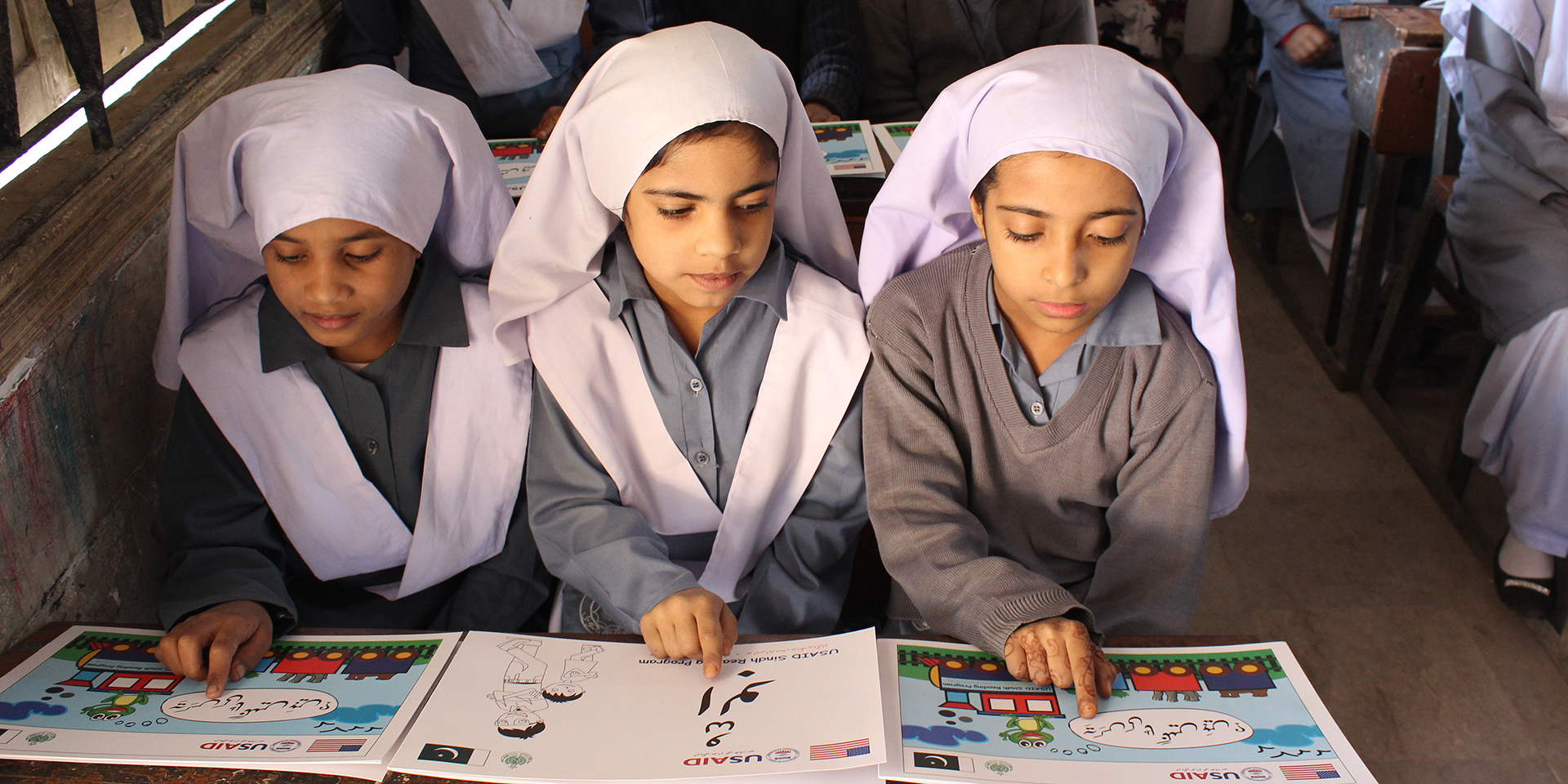 Image of three girls sitting at a school desk and pointing at words on individual learning materials marked 