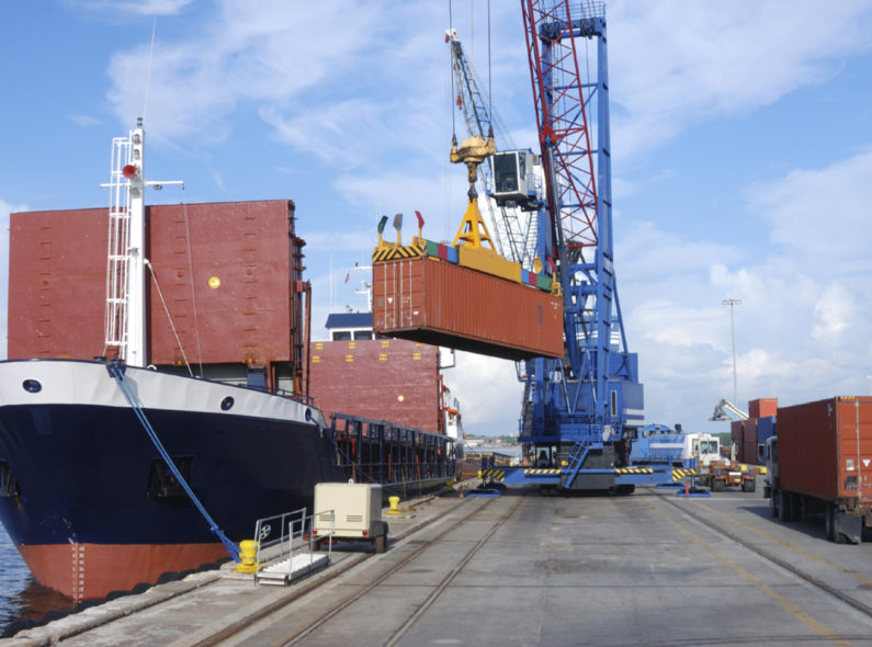 Image of a dock with a large shipping container being loaded onto a trading vessel by a blue crane.