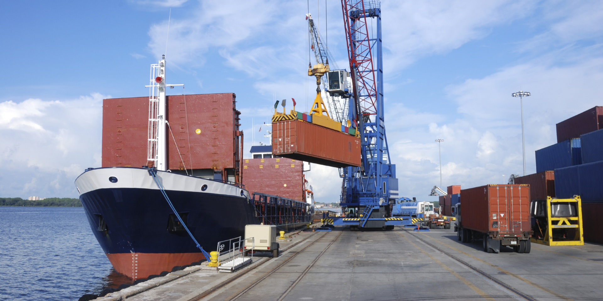 Image of a dock with a large shipping container being loaded onto a trading vessel by a blue crane.
