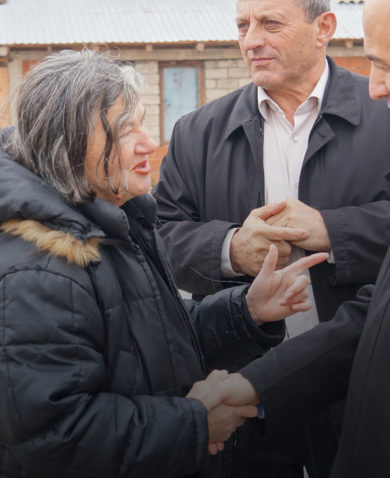 Image of a woman in a padded coat smiling and shaking hands with a man in an overcoat on a street.