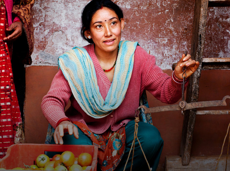 A woman kneeling in front of a wall holding a scale in her right hand and reaching for tomatoes in a red basket with her left hand.