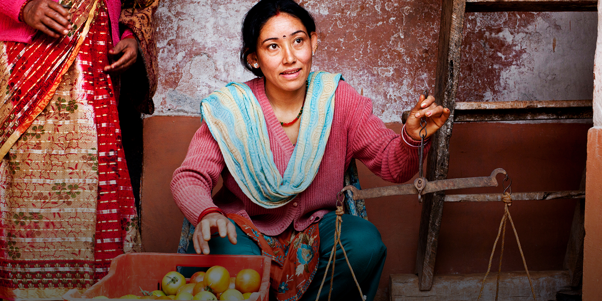 A woman kneeling in front of a wall holding a scale in her right hand and reaching for tomatoes in a red basket with her left hand.