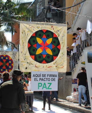 A street fair with several colorful banners decorating the road. The closest banner reads "Aqui se firma el pacto de PAZ." Several people are walking on each side of the road and viewing exhibits.