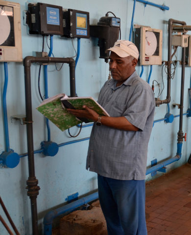 A man reads a notebook in a large room filled with pumps, valves, and large blue pipes.