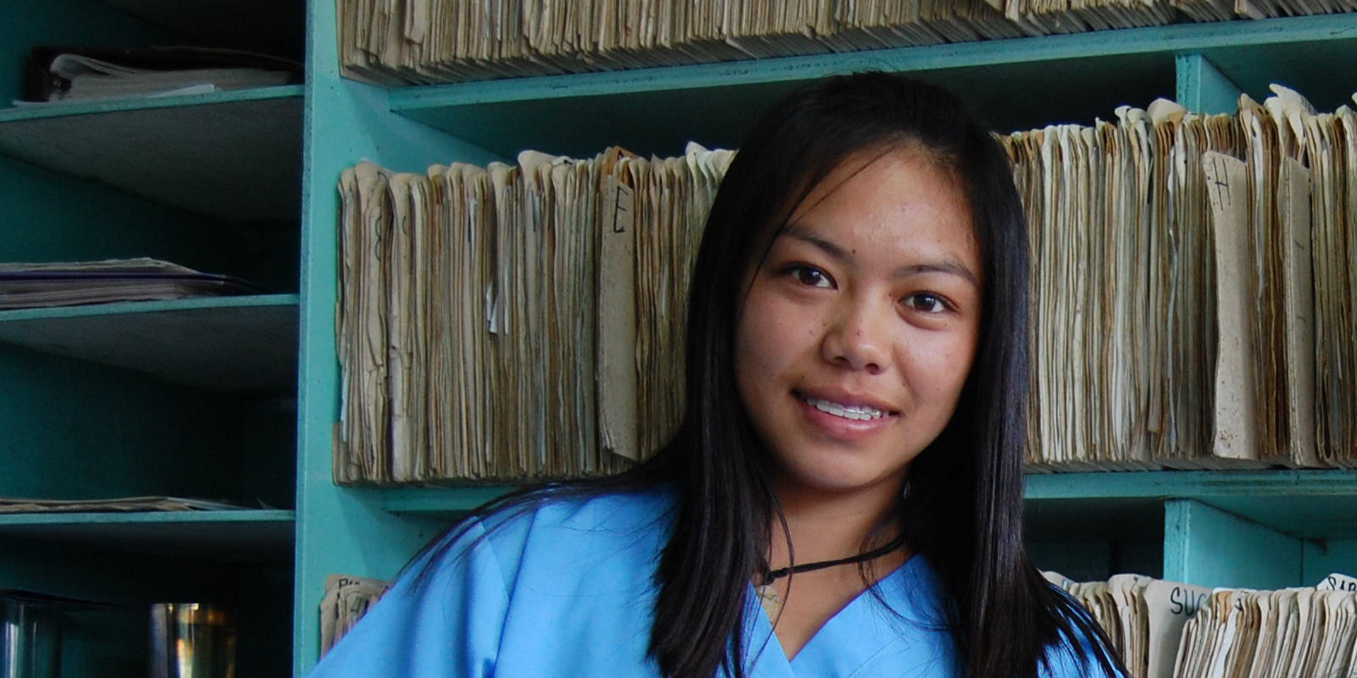 Image of a young female health care worker smiling in front of a shelf filled with manilla files.
