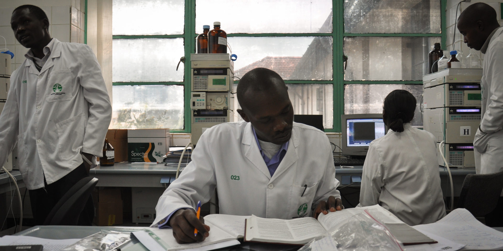 Several people in lab coats working amongst medical equipment. One in the center is reading through a large book and taking notes.