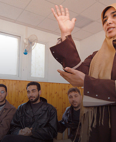 A woman stands and communicates with sign language as several men sit and watch in the background.