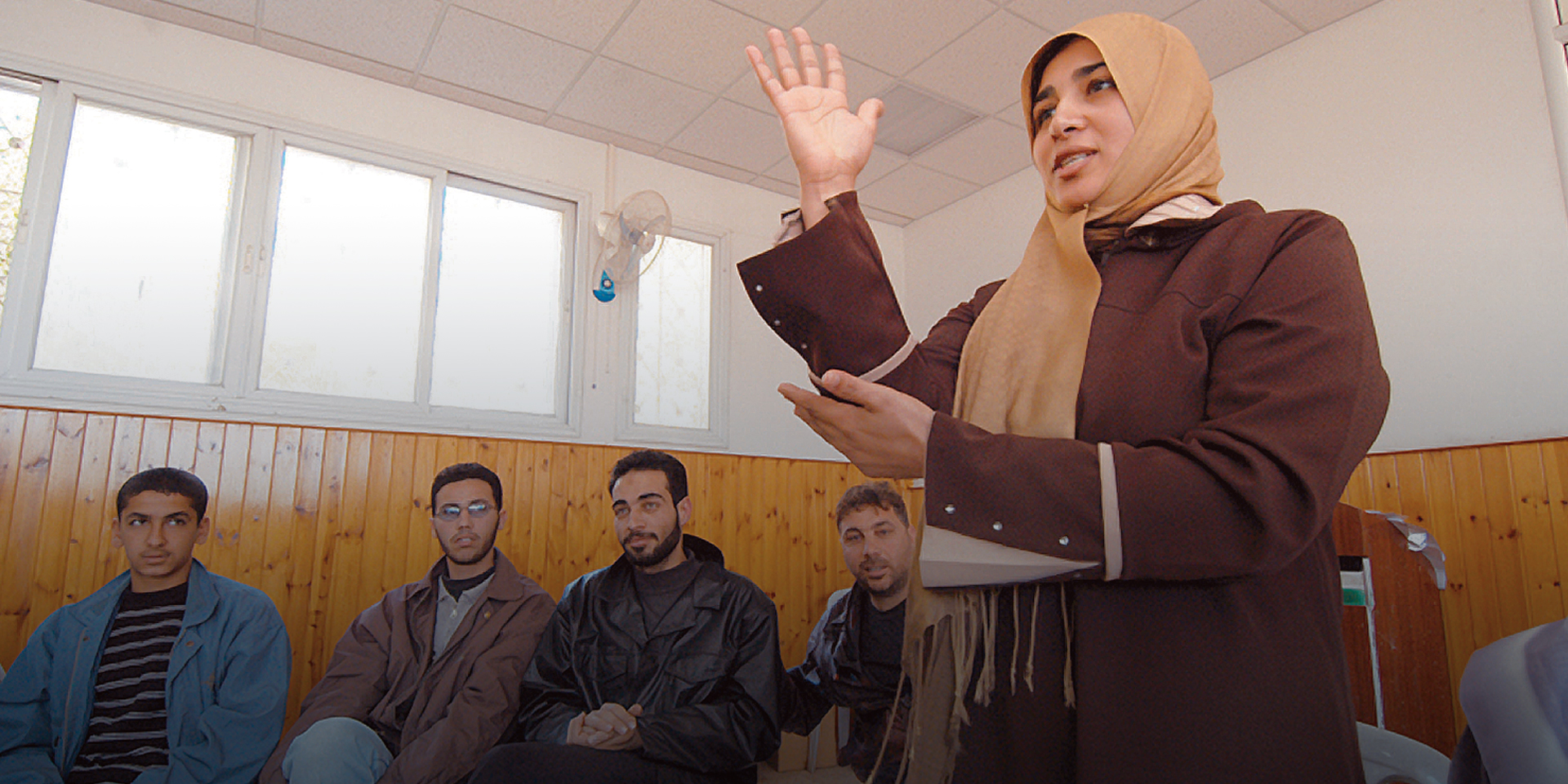 A woman stands and communicates with sign language as several men sit and watch in the background.