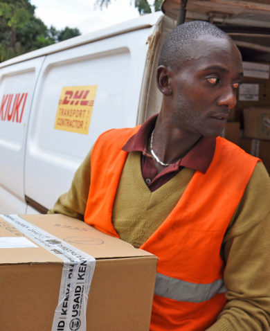 A man carrying a box labeled "USAID | Kenya" out of a truck marked "DHL."