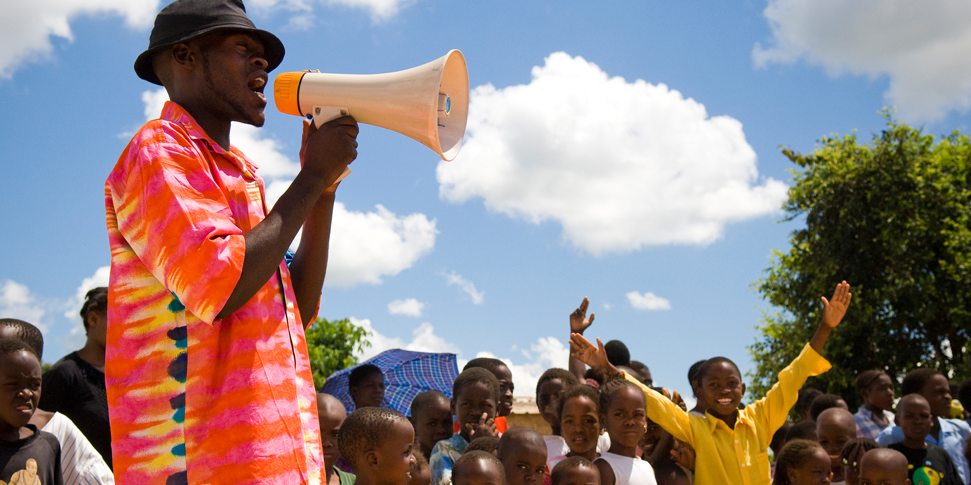 A man in a brightly colored shirt speaking into a megaphone as several children stand beside him.