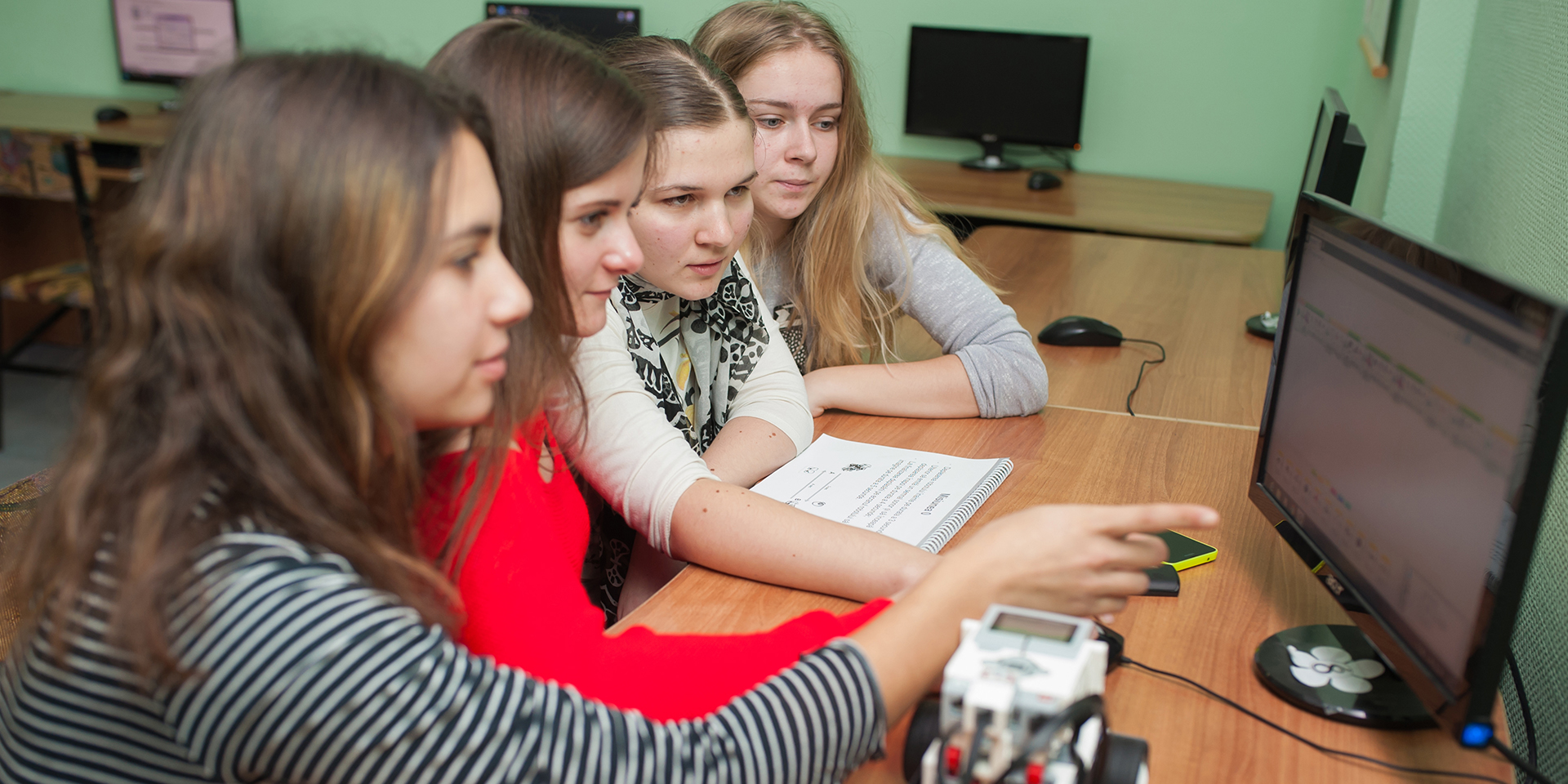 A group of four girls sitting in front of a computer monitor as one points out something on the screen.