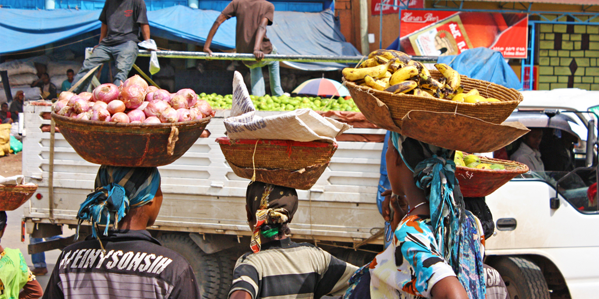 several women holding fruits and vegetables in baskets on their heads watching as a large white truck drives past.