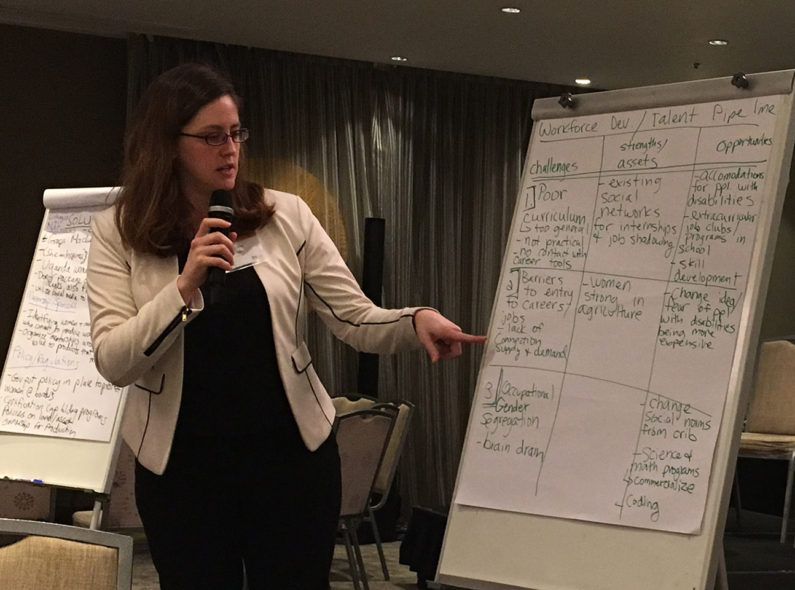 Image of two women giving a presentation as one points to a whiteboard listing several items under the headers "Challenges," "Strengths/Assets," and "Opportunities."