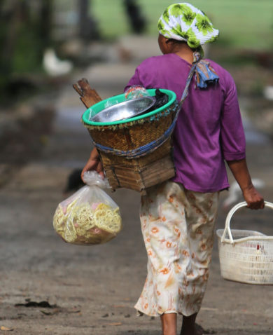 Image of a woman walking away on a paved road carrying several baskets and a bag.