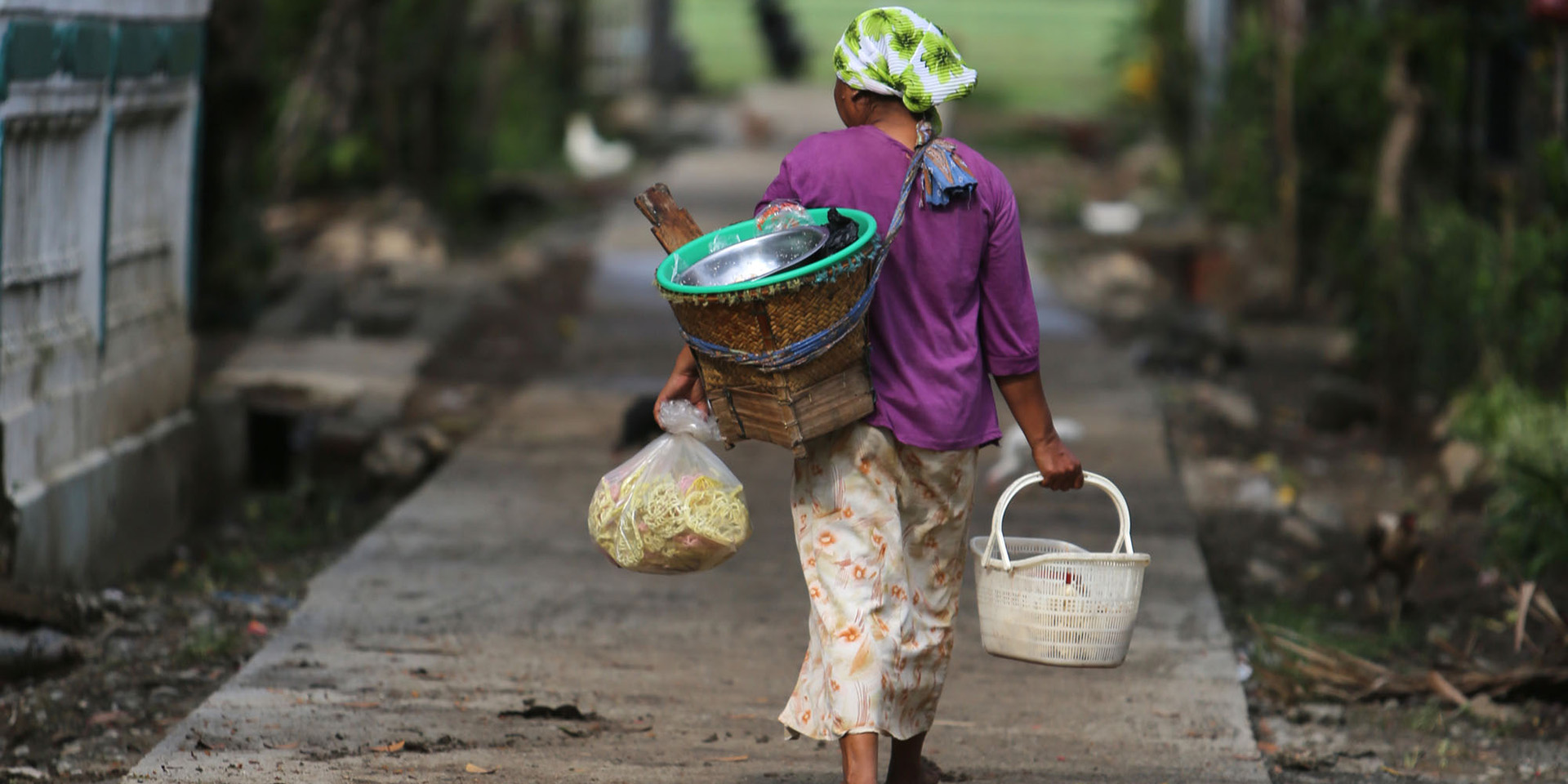 Image of a woman walking away on a paved road carrying several baskets and a bag.