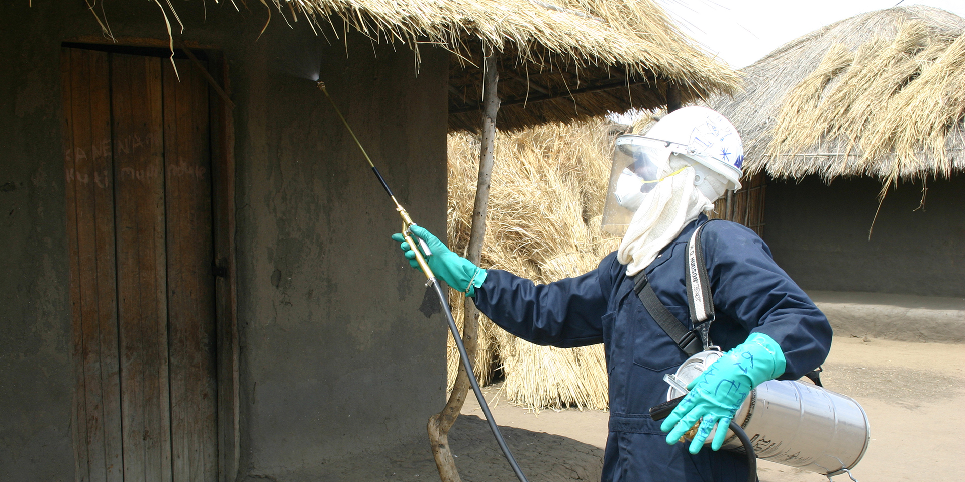 Image of a man wearing protective equipment spraying pesticides on a hut.