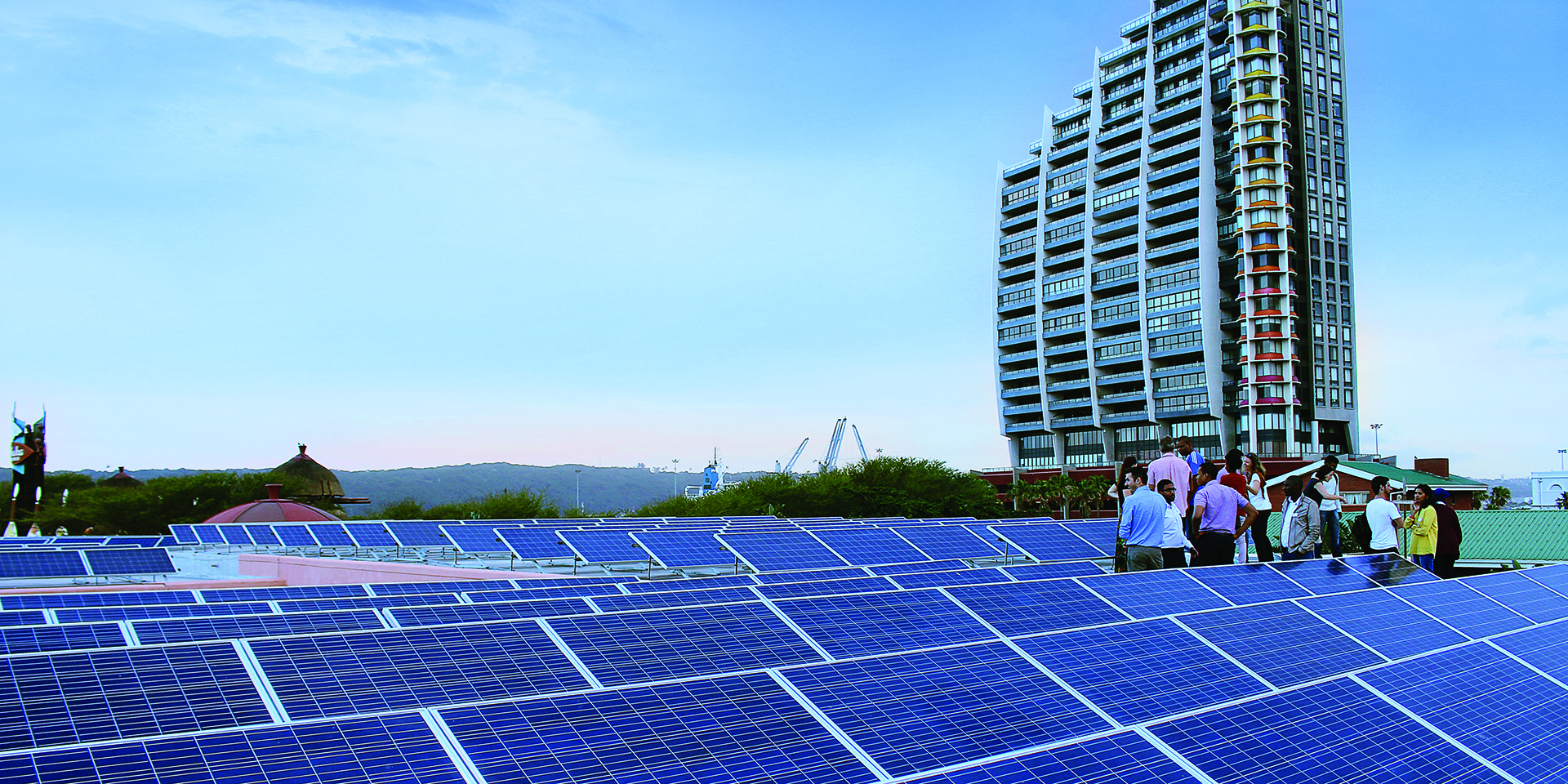 Image of a large group of people standing in the middle of a solar array. A large building and cranes can be seen in the distance.