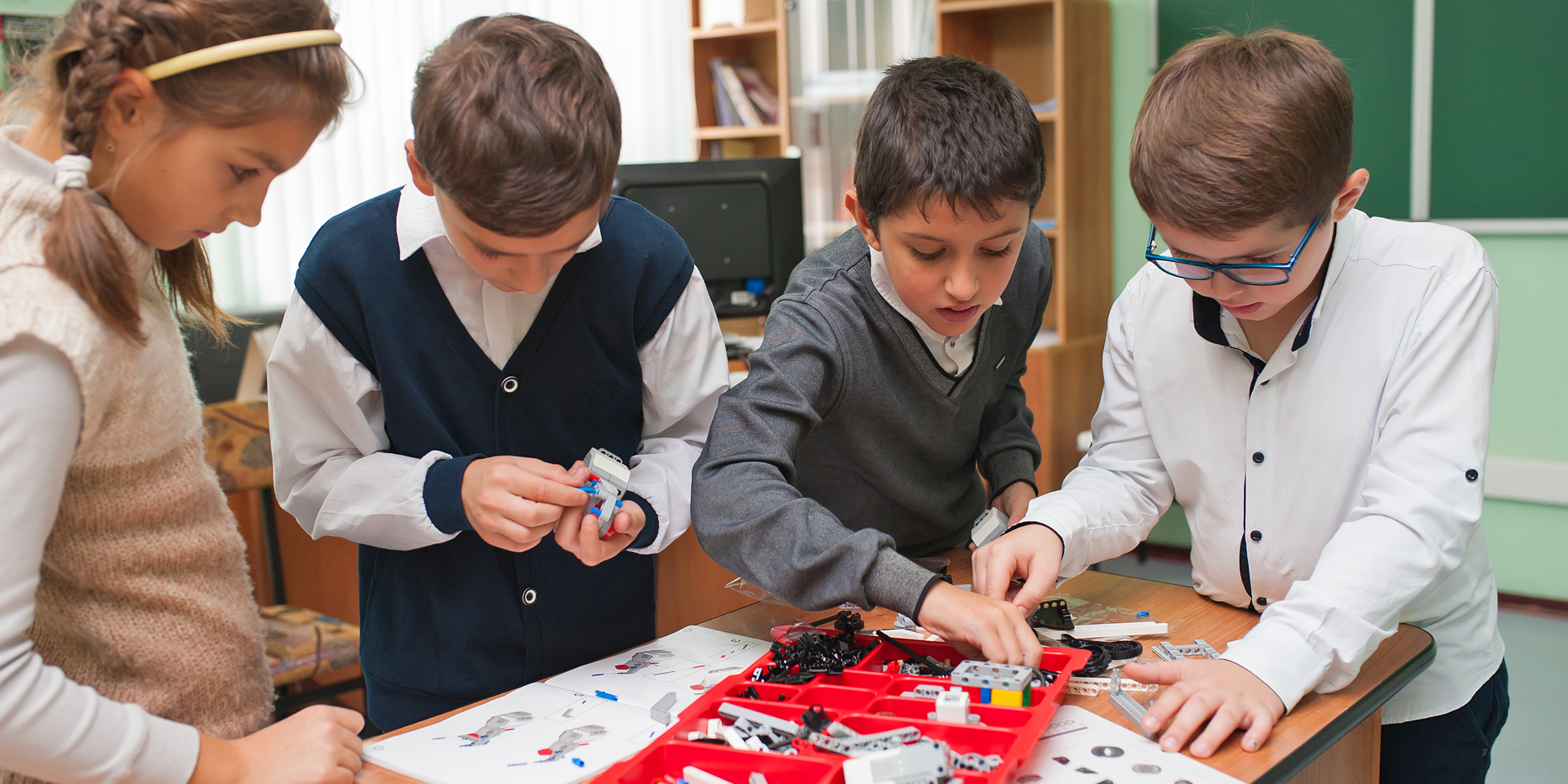 Four children putting together a device using Lego-like parts in a red tray.