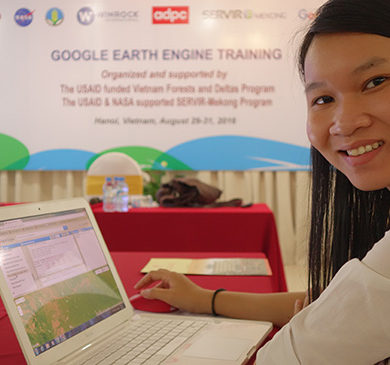 Image of a woman smiling at the camera as she sits in front of a laptop. In the background hangs a poster that says "Google Earth Engine Training."
