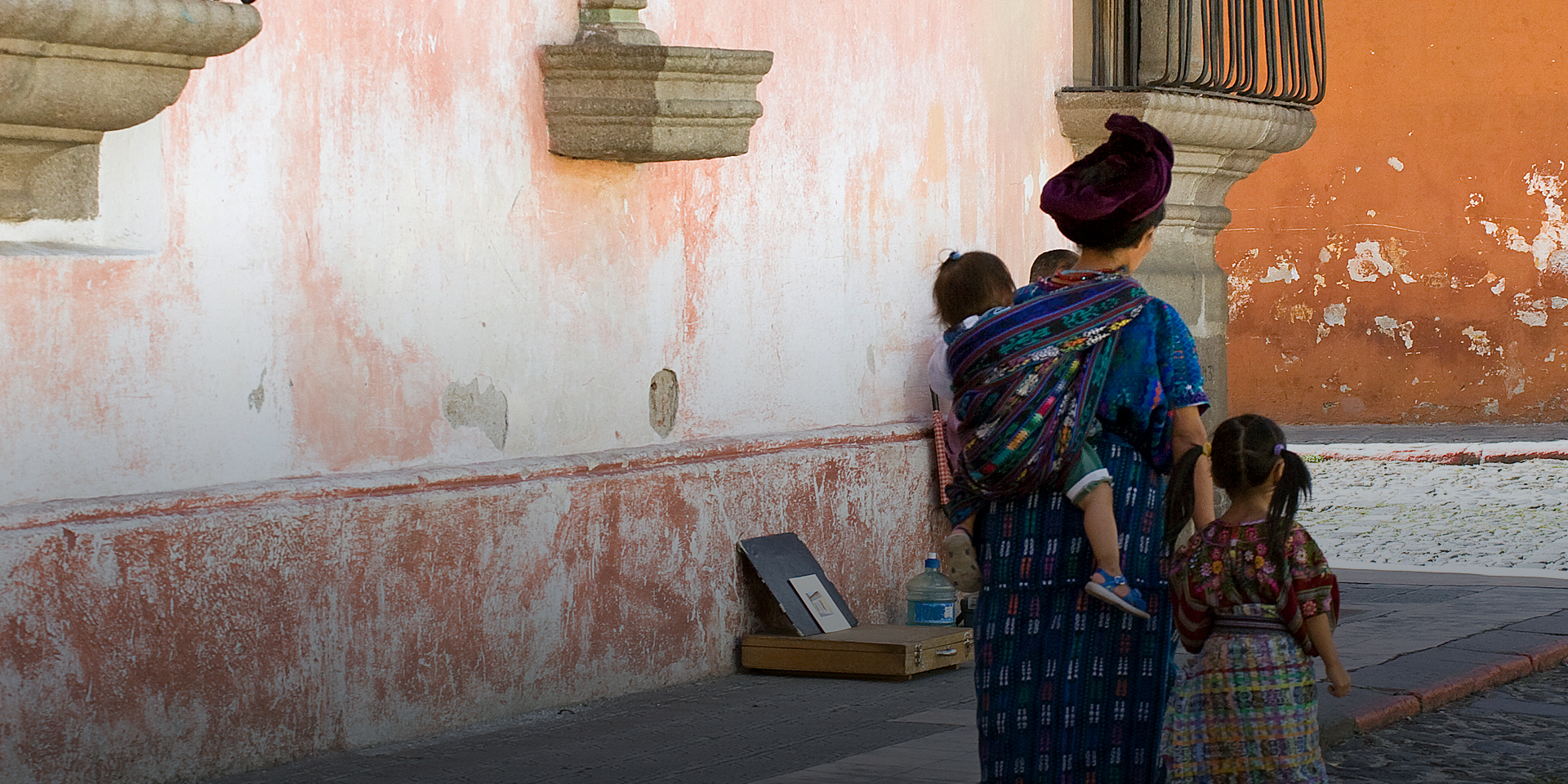 A mother walks past a red-tinged wall with a baby wrapped in a blanket on her back and a toddler walking beside her.