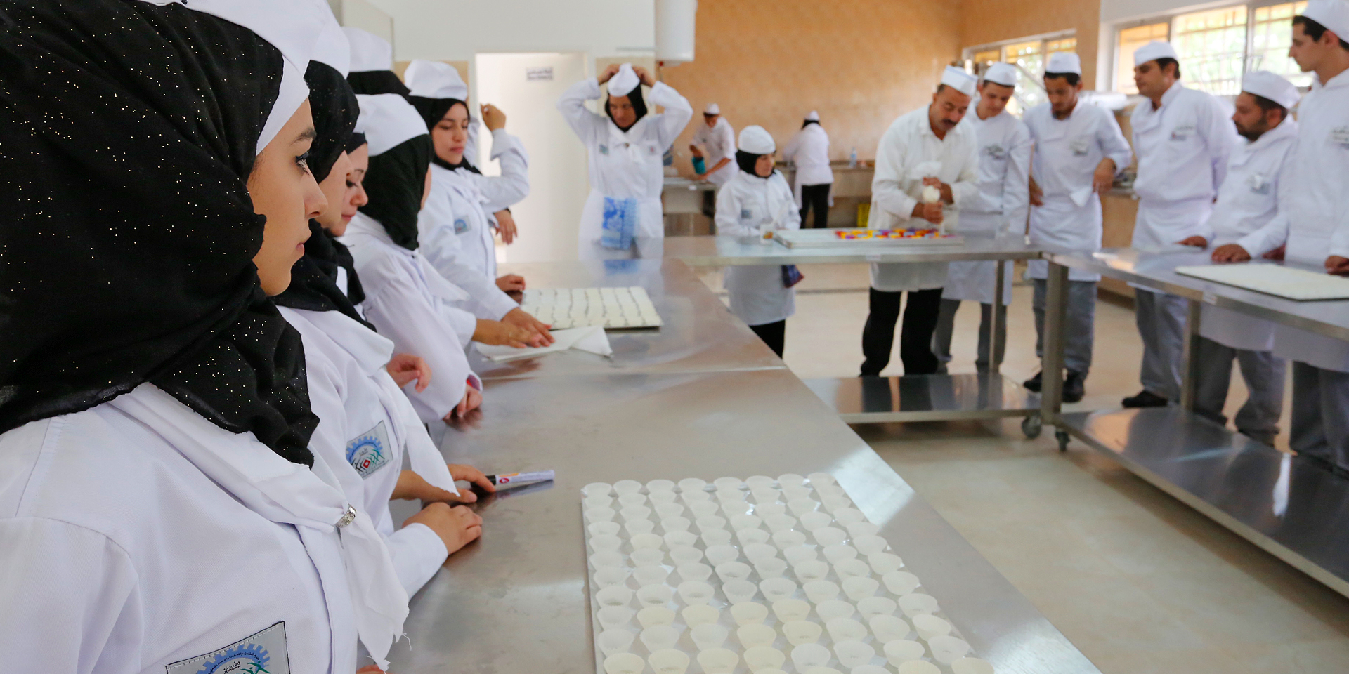 A room filled with cooks standing around silver tables with paper cups on a cookie sheet sitting upon them. A chef stands in the center demonstrating a preparation method.