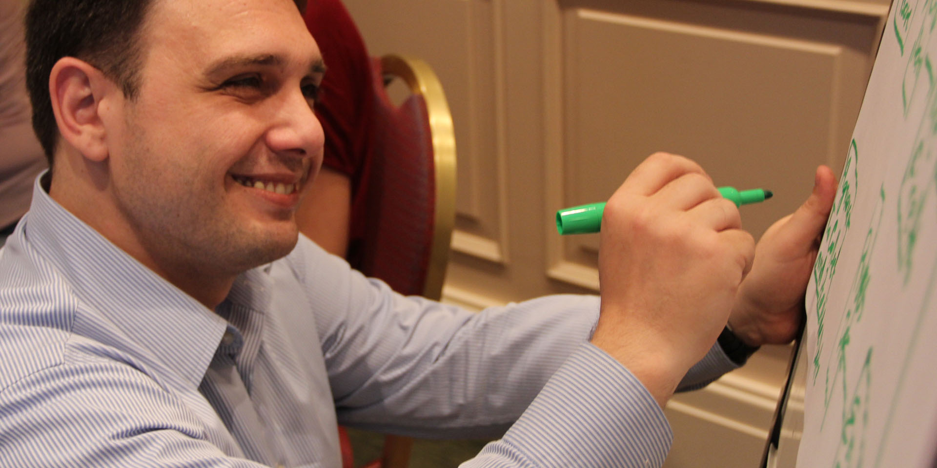 A smiling man writing on a whiteboard with a green marker.