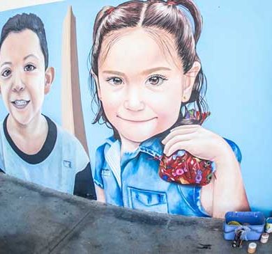 A mural being painted on a wall showing two smiling children with a monument in the background and a cheerful dog with a red and white leash.