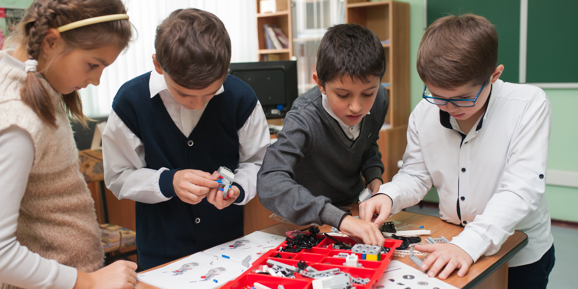 Four children putting together a small robot with parts from a red container sitting on a table.
