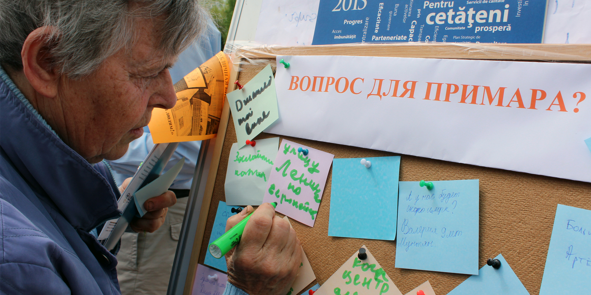 A man outside writing on a sticky note pinned to a corkboard.