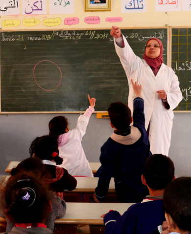 A schoolroom filled with children at desks with several raising their hands. A teacher standing in front of a chalkboard points to one of the students raising their hands in the back of the room.
