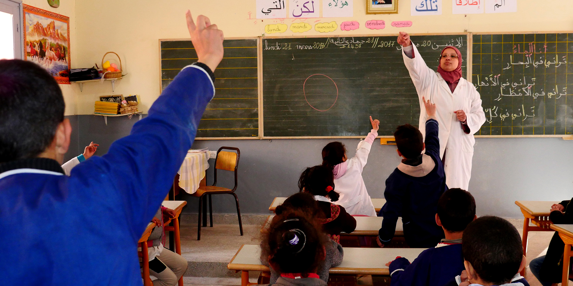 A schoolroom filled with children at desks with several raising their hands. A teacher standing in front of a chalkboard points to one of the students raising their hands in the back of the room.