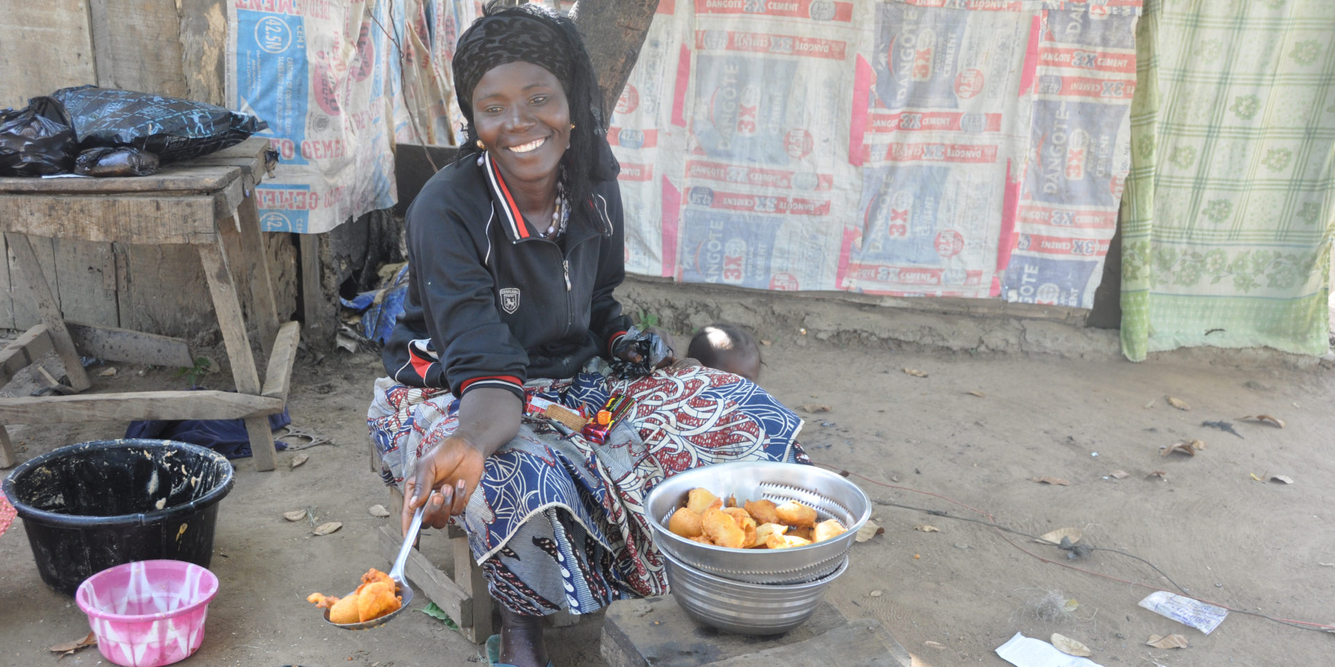 Image of a smiling woman sitting beside a bowl of fried food and offering some to someone off-camera with a large spoon.