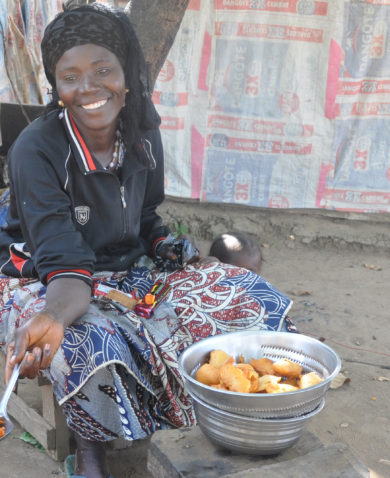 Image of a smiling woman sitting beside a bowl of fried food and offering some to someone off-camera with a large spoon.