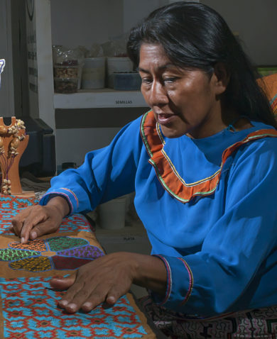 Two women sitting across from each other at a table. One watches as the other points to a bright quilt on the table and explains her process.