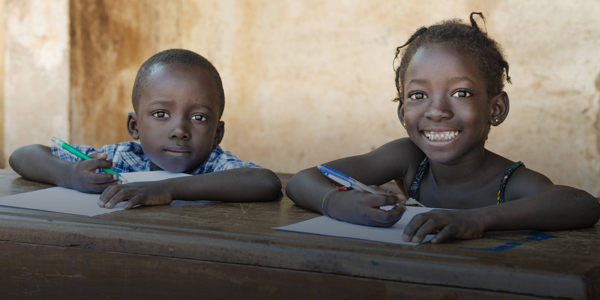 Two children smiling while sitting at a long bench and writing with pens on individual pieces of paper.