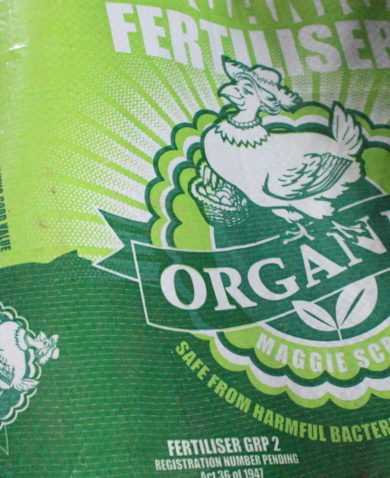 A close-up image of a green bag of fertilizer called "Organic; Maggie Scratcher," with a cartoon depiction of a smiling chicken.