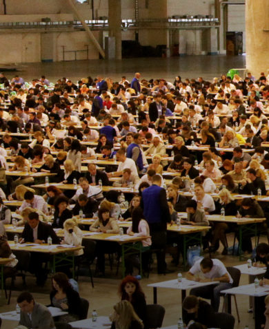 The interior of a large building filled with hundreds of people sitting at tables.