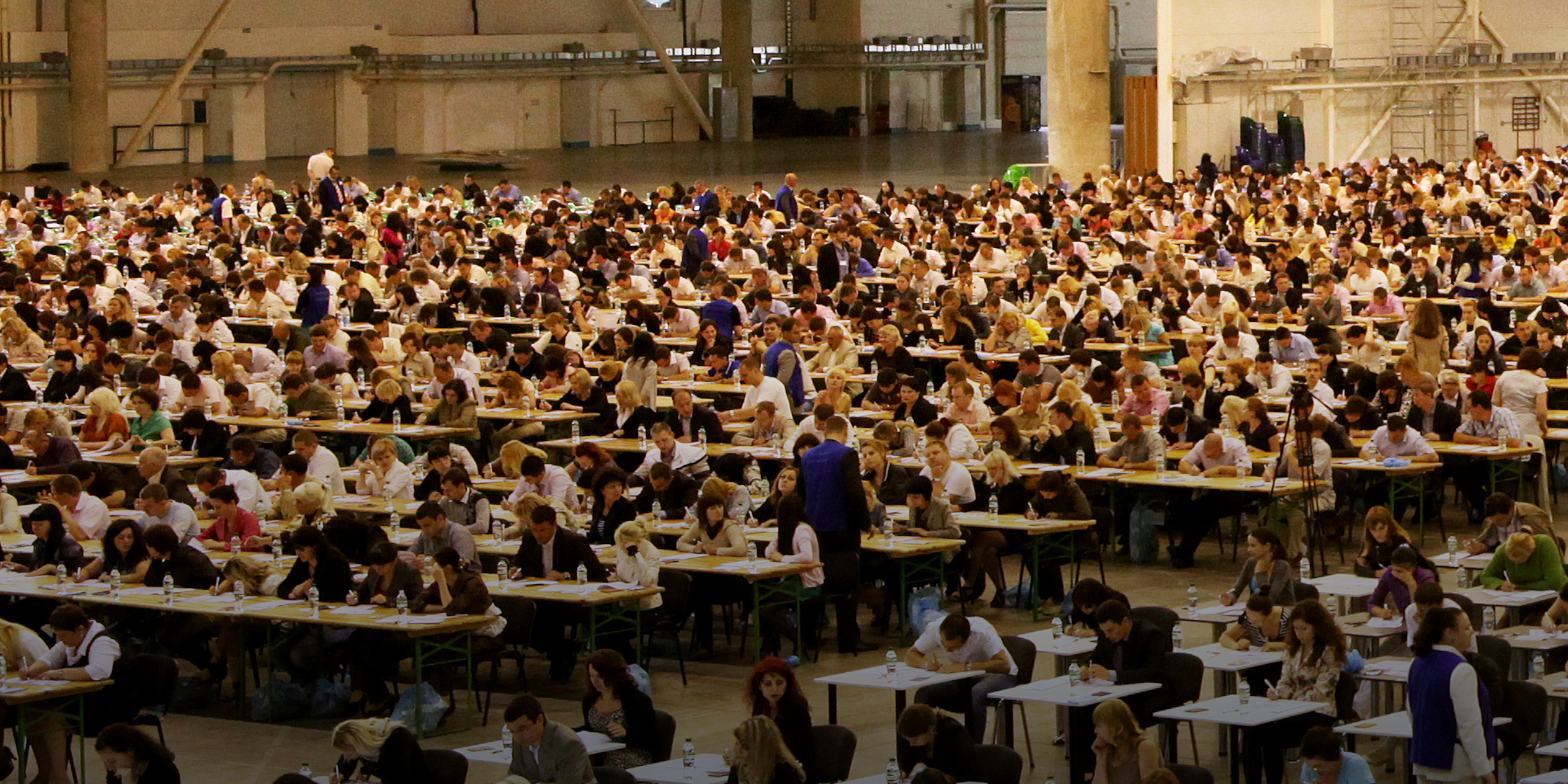 The interior of a large building filled with hundreds of people sitting at tables.