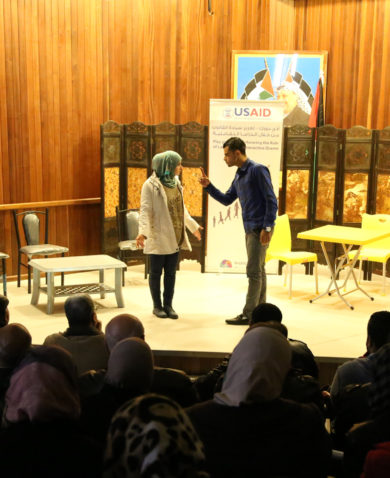 A stage performance with two people acting for an audience of people. A sign marked "USAID" is in the background.
