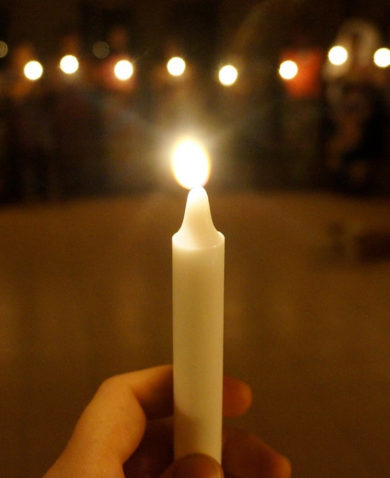 A close-up image of a burning candle being held by a hand. Several people holding candles can be seen standing in a circle in the background.