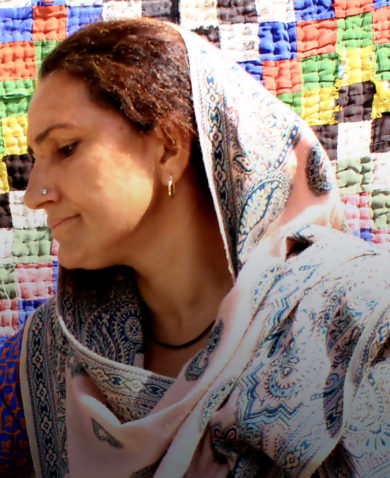A woman looking off-camera and sitting in front of a large quilt composed of multi-colored squares.