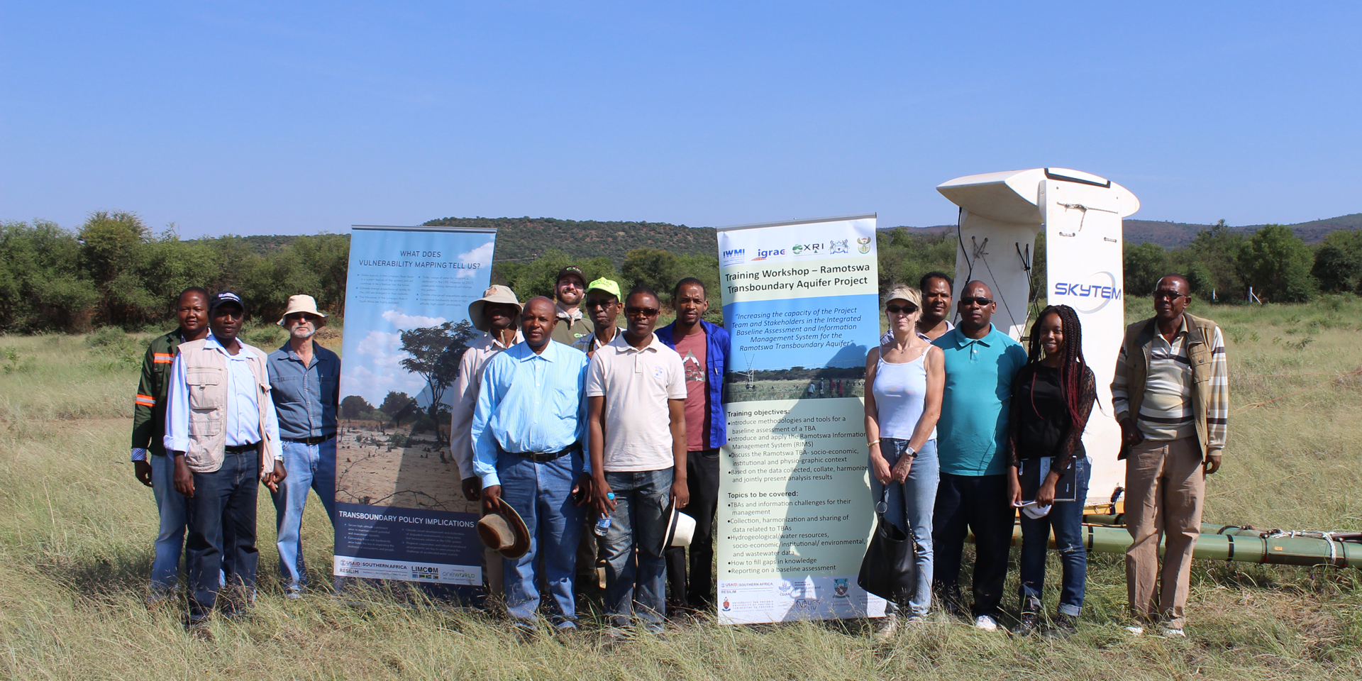 Image of several people in a field posing beside several posters for a training workshop. Behind them is a large drone marked 