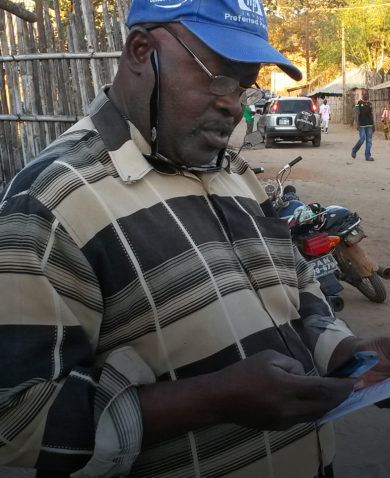 A man standing on a street corner typing into a cellphone while looking at a sheet of paper.