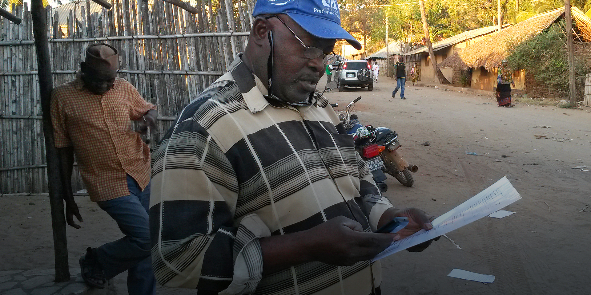 A man standing on a street corner typing into a cellphone while looking at a sheet of paper.